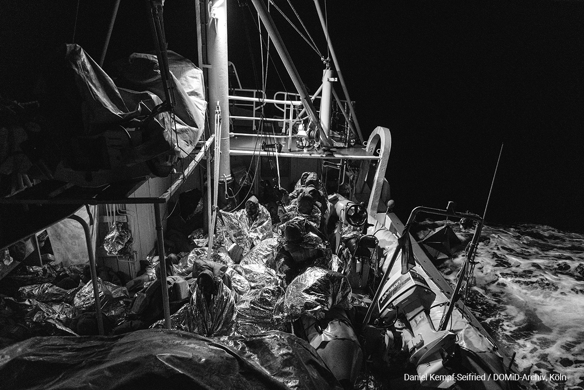 Das Foto ist bei einer Rettungsaktion im April 2017 entstanden. Daniel Kempf-Seifried war mit an Bord: „Ich war mit einem Rettungsboot der Nichtregierungsorganisation Sea-Eye an der libyschen Küste unterwegs um Schiffbrüchige mit Schwimmwesten auszustatten, so der Plan. Doch es kam alles ganz anders als gedacht. […] Die Iuventa und die Sea-Eye versuchten die Menschen so gut es ging abzubergen und auf Rettungsinseln als auch an Deck der Rettungsboote zu bringen. Nach einigen Stunden erreichte der Bundeswehr Tender RHEIN das Szenario und half uns mit zwei RIB Booten die verbliebenen Menschen zu bergen.“  Foto: Daniel Kempf-Seifried / DOMiD-Archiv, Köln